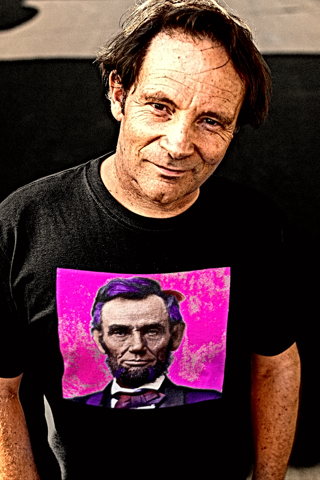 Tough middle aged Whtie guy earing a basic black t-shirt with a stylized photo of Abraham Lincoln: American Punk From his Second Inaugural Address, delivered near the end of the Civil War, Lincoln’s words capture the nation’s moral high point amid its deepest wound. Great gift for American radicals and history buffs. Bella+Canvas 3001 Basic T-Shirt #EUTC

