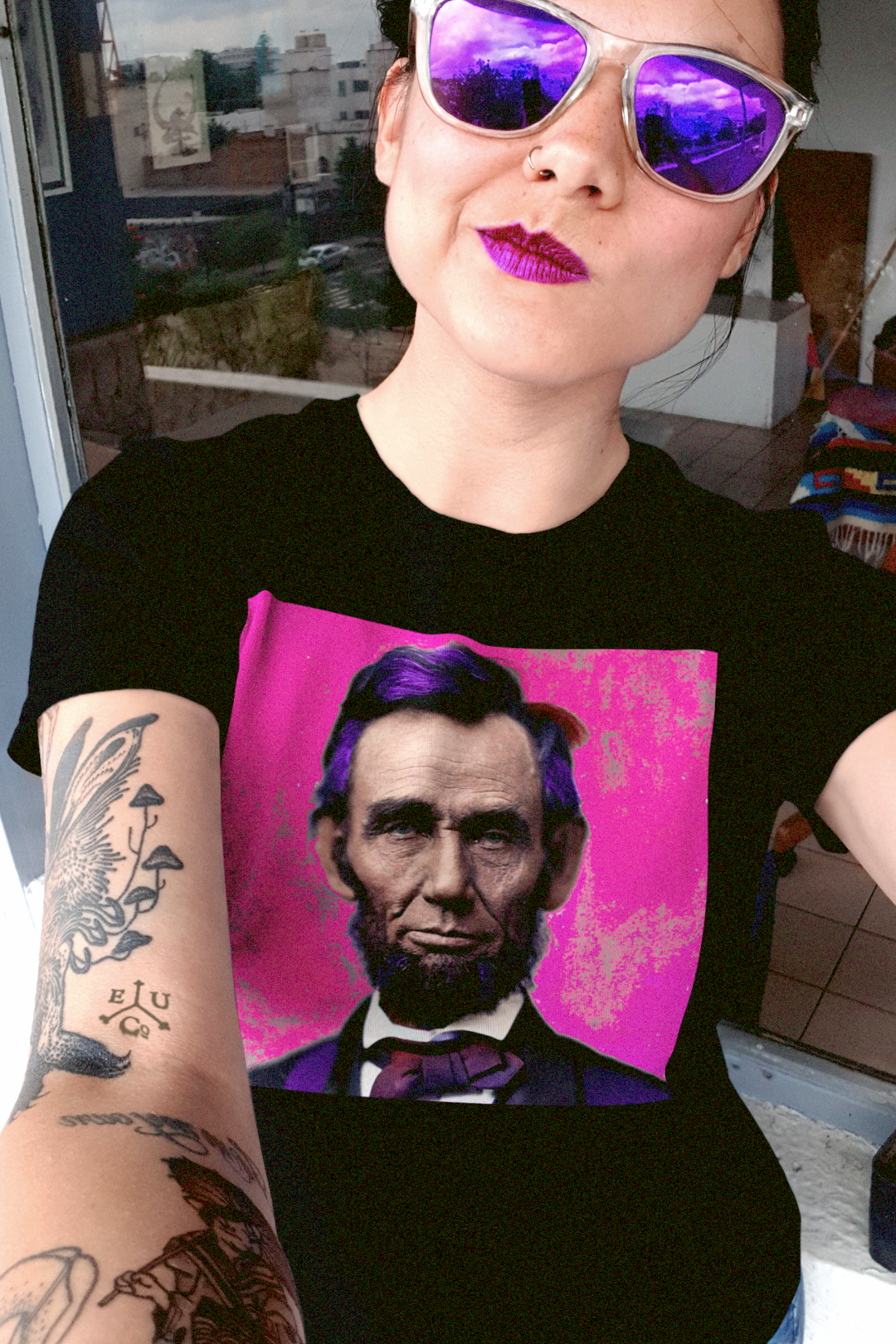 Tattooed woman with purple reflector sunglasses wearing a Black relaxed fit women's tee depicting Abraham Lincoln: American Punk. Technicolorization against a hot pink background. Lincoln wasn’t a saint dropped from the heavens—he was a messy, pragmatic politician shaped by the rough-and-tumble Free Soil movement and the hard edges of frontier republicanism. Perfect gift for the radical history buff of the American Punk in your life. Bella+Canvas Women’s Relaxed Fit Tee 6400 #EUTC
