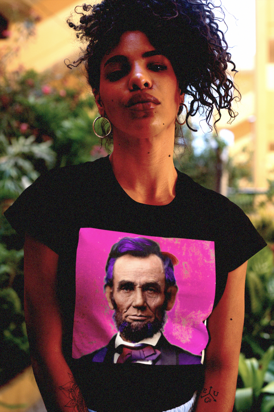 Serious but funky Black woman with kinky hair wearing a Black relaxed fit women's tee depicting Abraham Lincoln: American Punk. Technicolorization against a hot pink background. Lincoln wasn’t a saint dropped from the heavens—he was a messy, pragmatic politician shaped by the rough-and-tumble Free Soil movement and the hard edges of frontier republicanism. Perfect gift for the radical history buff of the American Punk in your life. Bella+Canvas Women’s Relaxed Fit Tee 6400 #EUTC
