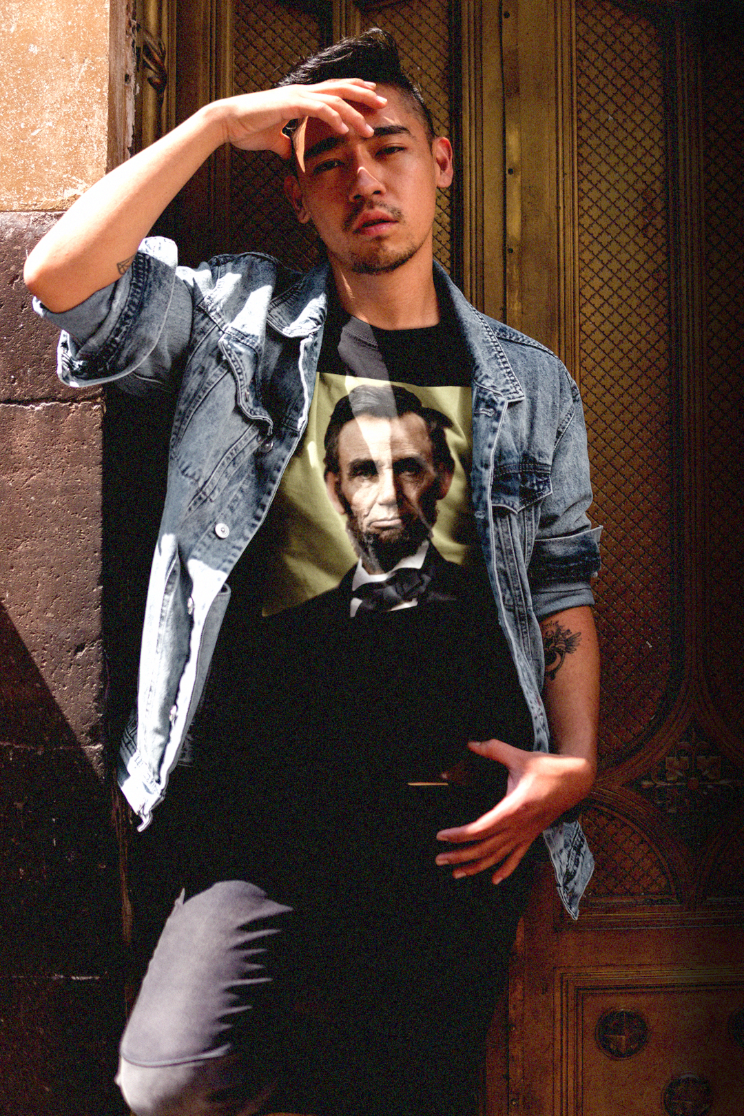 Asian guy with jean jacket in front of old carved wooden door wearing a Basic Black T-Shirt with khaki colorized portrait of Abraham Lincoln. From his Second Inaugural Address, delivered near the end of the Civil War, Lincoln’s words capture the nation’s moral high point amid its deepest wound. Great gift for American radicals and history buffs. Bella+Canvas 3001 Basic T-Shirt #EUTC
