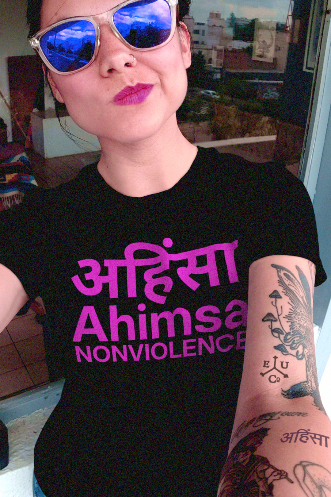 Tattooed woman with blue reflective sunglasses in a black relaxed fit women's tee: AHIMSA — The Principle of Nonviolence
Mahatma Gandhi transformed Ahimsa अहिंसा into a political force, extending it from private virtue to public resistance. 
Photo shows fit and style of the tee shirt. Perfect gift for the activist and nonviolent protester in your life. Bella+Canvas 6400 Women’s Relaxed Tee #EUTC