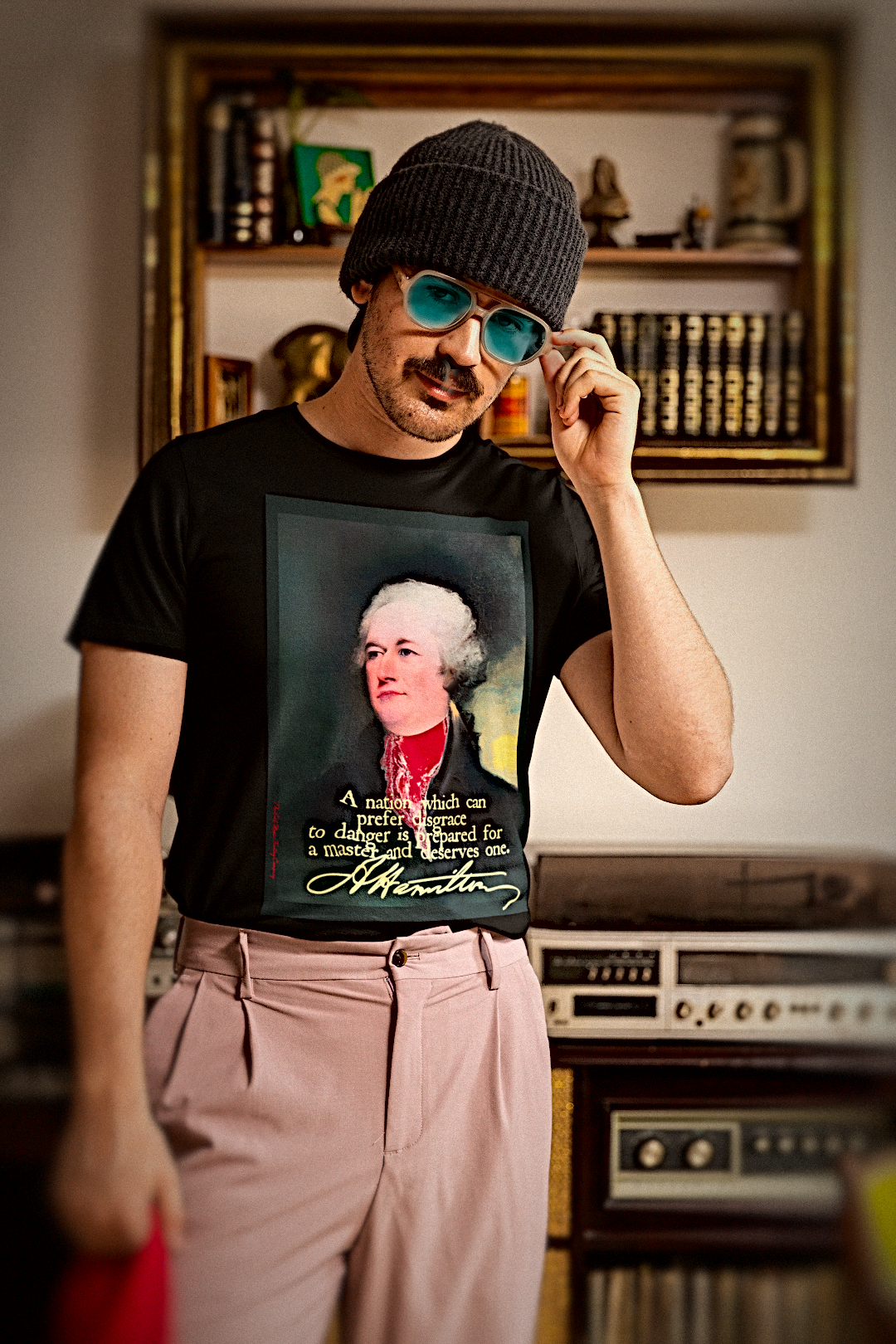 A man wearing a black graphic t-shirt with a portrait of Alexander Hamilton, styled with a black knit beanie, green-tinted sunglasses, and light pink trousers, standing in a vintage room with a record player and bookshelves.