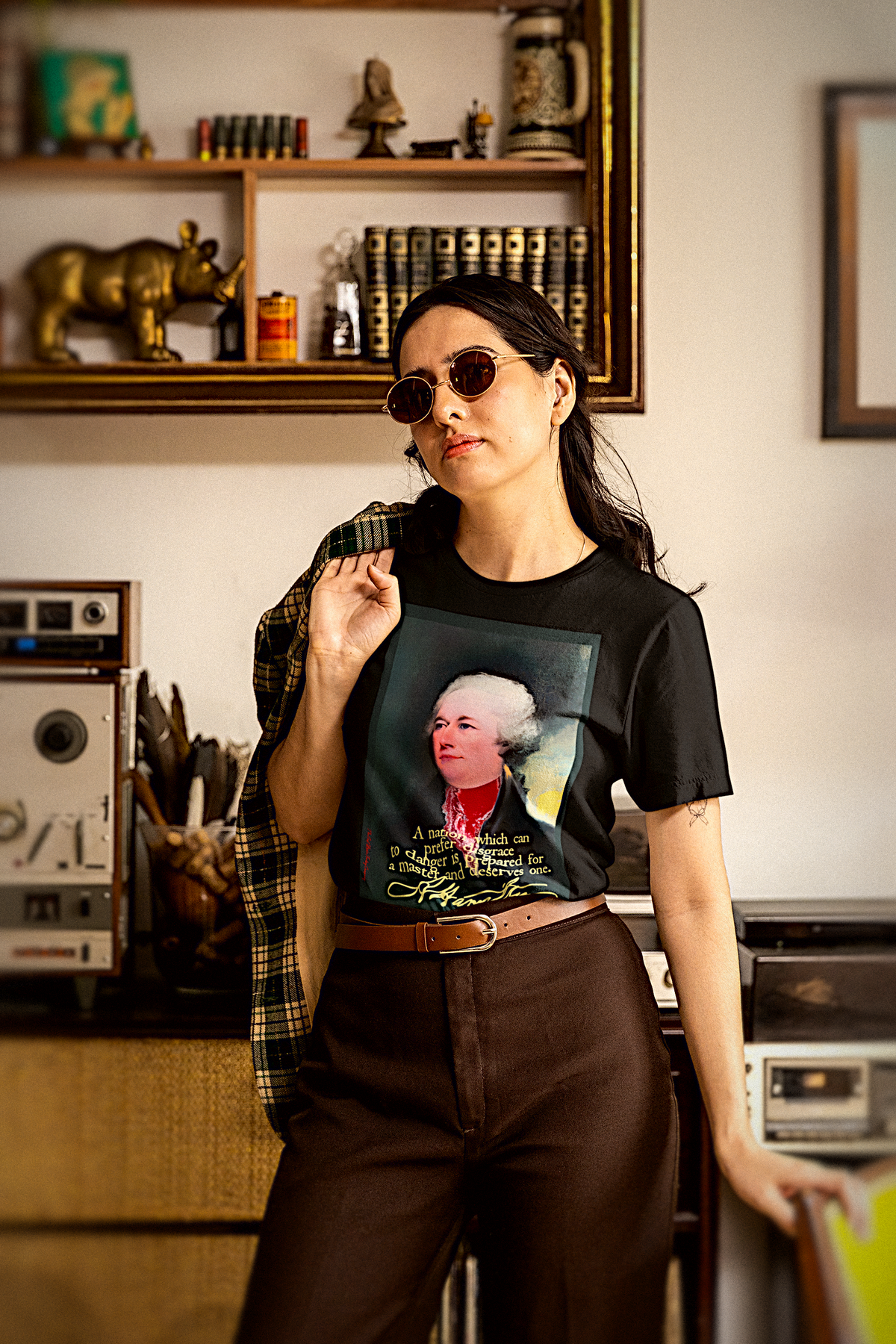 A woman wearing a black graphic t-shirt with a portrait of Alexander Hamilton, styled with brown trousers and a plaid shirt draped over her shoulder, standing in a vintage room with retro electronics and bookshelves.