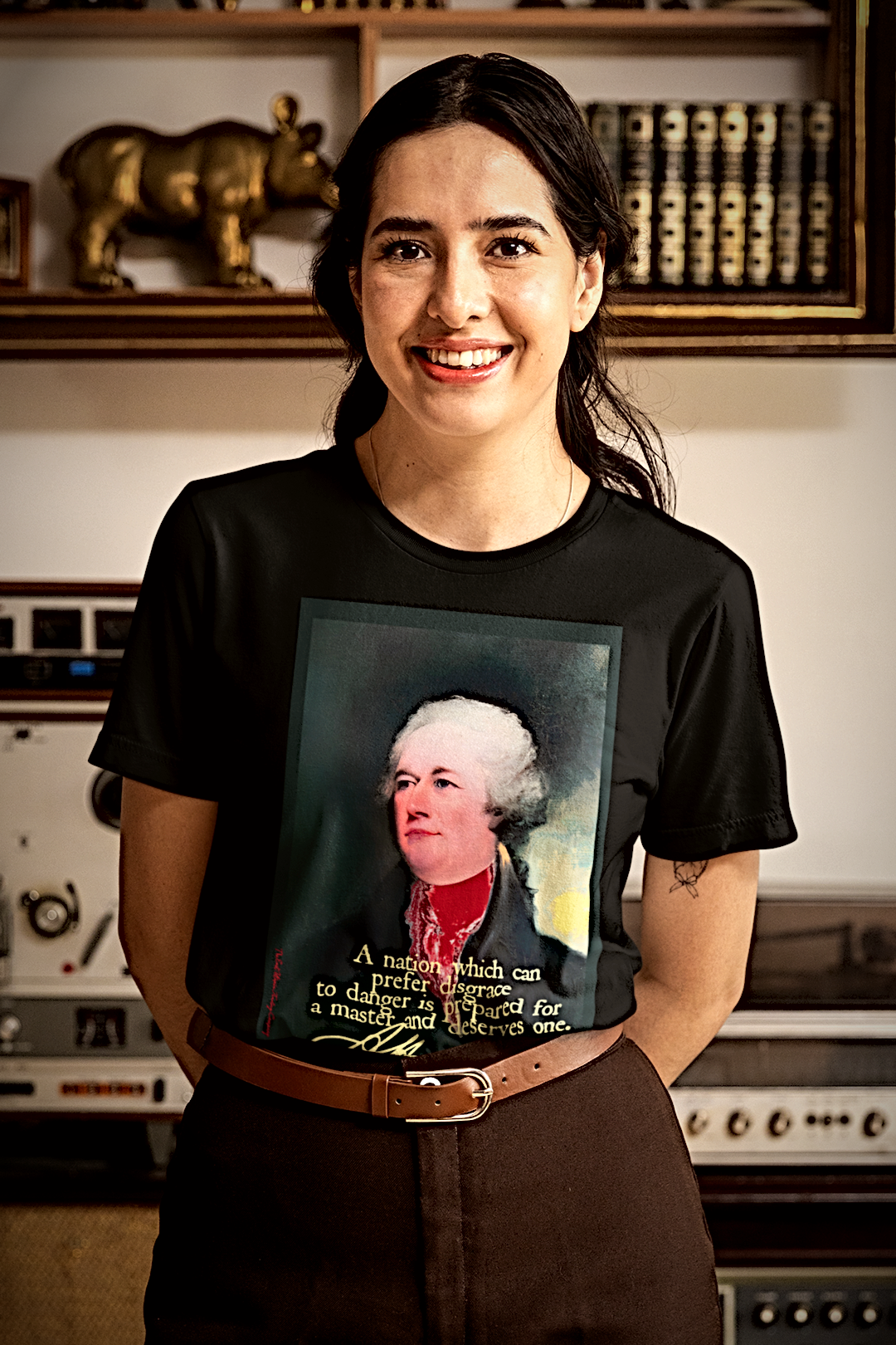A woman smiling while wearing a black graphic t-shirt featuring a portrait of Alexander Hamilton with his signature quote, set against a vintage audio equipment and bookshelf background.