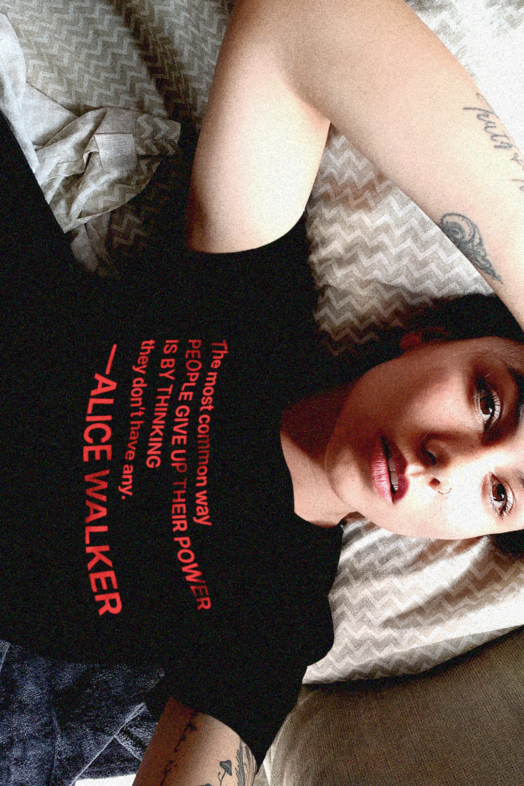Tattooed Asian American woman laying in bed wearing a Black Relaxed Fit Women's Tee with the Alice Walker quote “The most common way people give up their power is by thinking they don't have any.” —Poet, novelist, and activist Alice Walker (b. 1944) distills the essence of empowerment in this call to awareness. Great gift for your book club friend or the feminist activist in your life. Cotton Bella+Canvas 6400 #EUTC