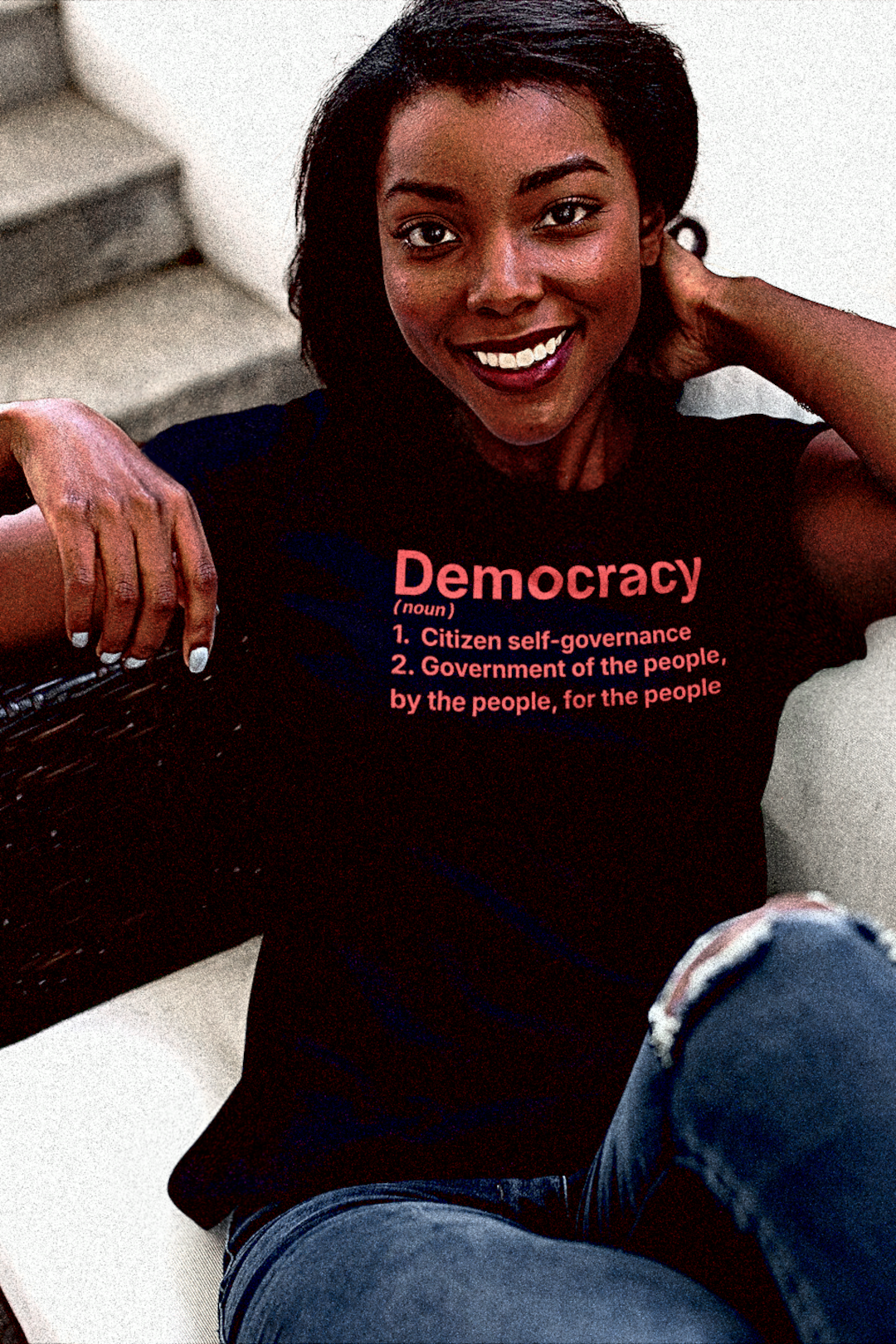 Trim Black woman sitting in rattan chair wearing a Quality Relaxed Fit Women's Tee with The Activist's Dictionary definition of DEMOCRACY(noun)  1. Citizen self-governance  2. Government of the people, by the people, for the people | Perfect gift for the democracy activist in your life. Quality Bella+Canvas 6400