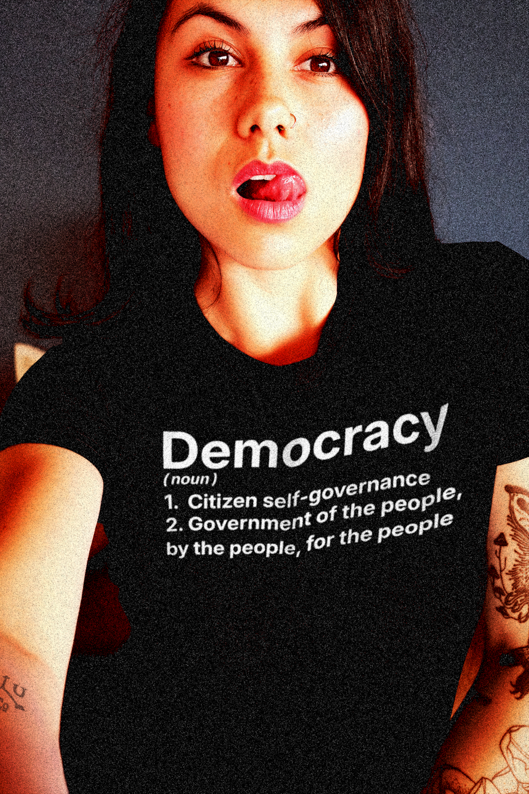 Tattooed Asian American woman sticking out her tongue wearing a Quality Relaxed Fit Women's Tee with The Activist's Dictionary definition of DEMOCRACY(noun)  1. Citizen self-governance  2. Government of the people, by the people, for the people | Perfect gift for the democracy activist in your life. Quality Bella+Canvas 6400