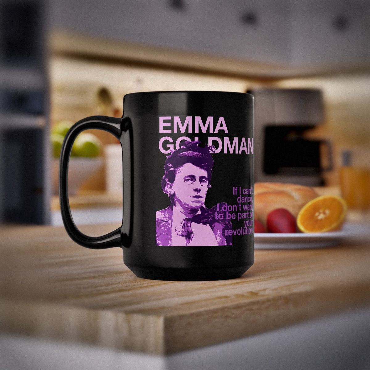 A black ceramic mug with a purple graphic print of Emma Goldman and text, set on a wooden countertop in a blurred kitchen environment.