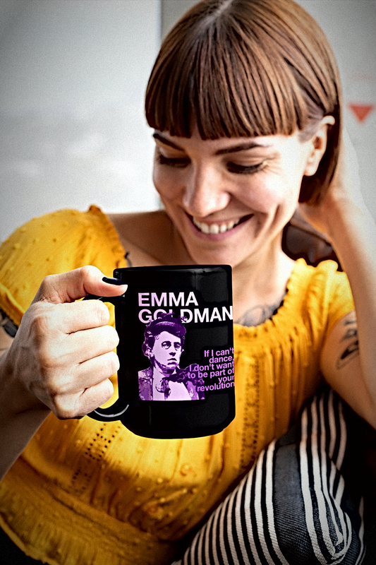 A woman in a yellow top holds up a black ceramic mug featuring a purple and white graphic of Emma Goldman with the text 'EMMA GOLDMAN' and the "If I can't dance" quote. The background is a soft-focus indoor setting.