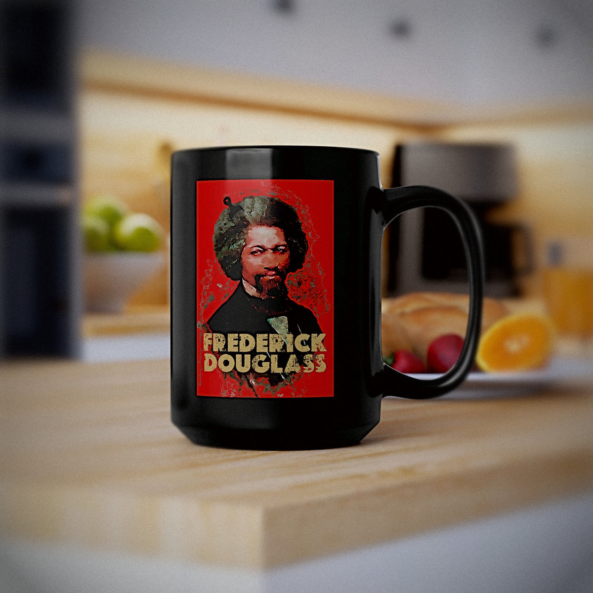 A black ceramic mug with a printed graphic of Frederick Douglass on a red background, sitting on a wooden countertop in a blurred kitchen setting.