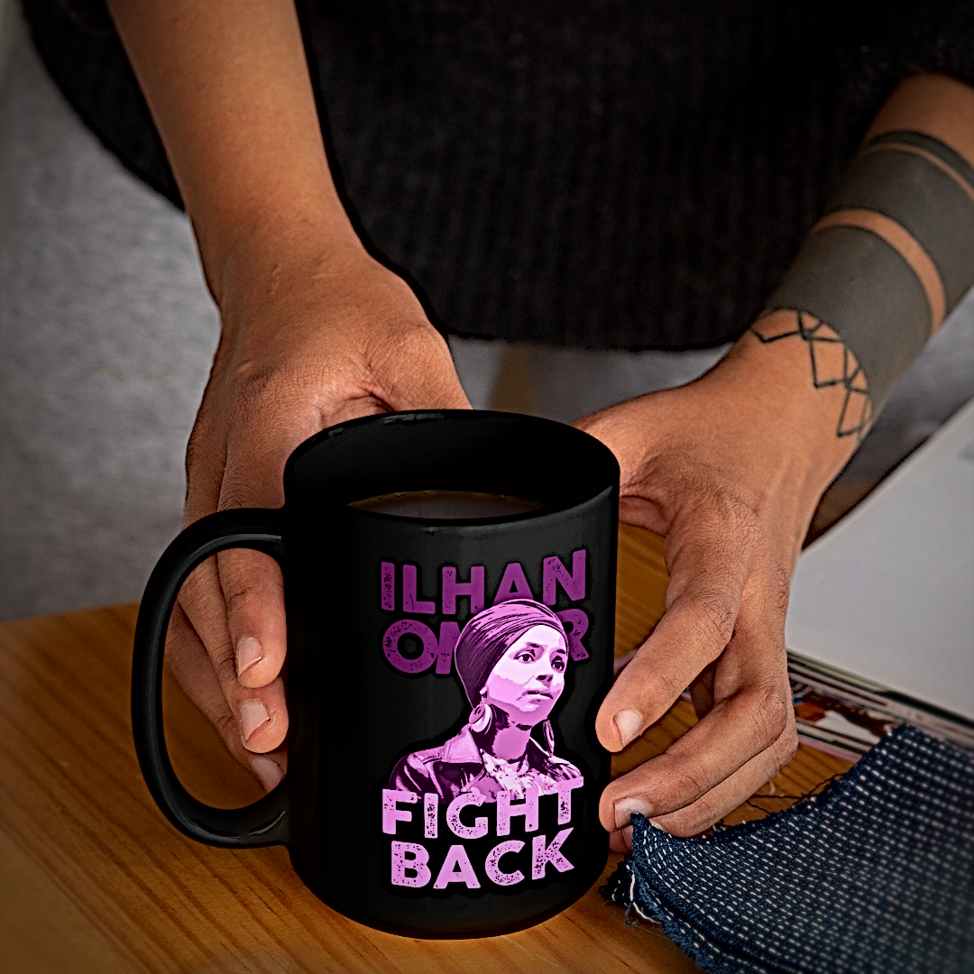 A person holds a black ceramic mug with a purple graphic print featuring the text 'ILHAN OMAR' and 'FIGHT BACK' alongside an illustrated portrait. The mug is shown on a wooden table with a gray background.