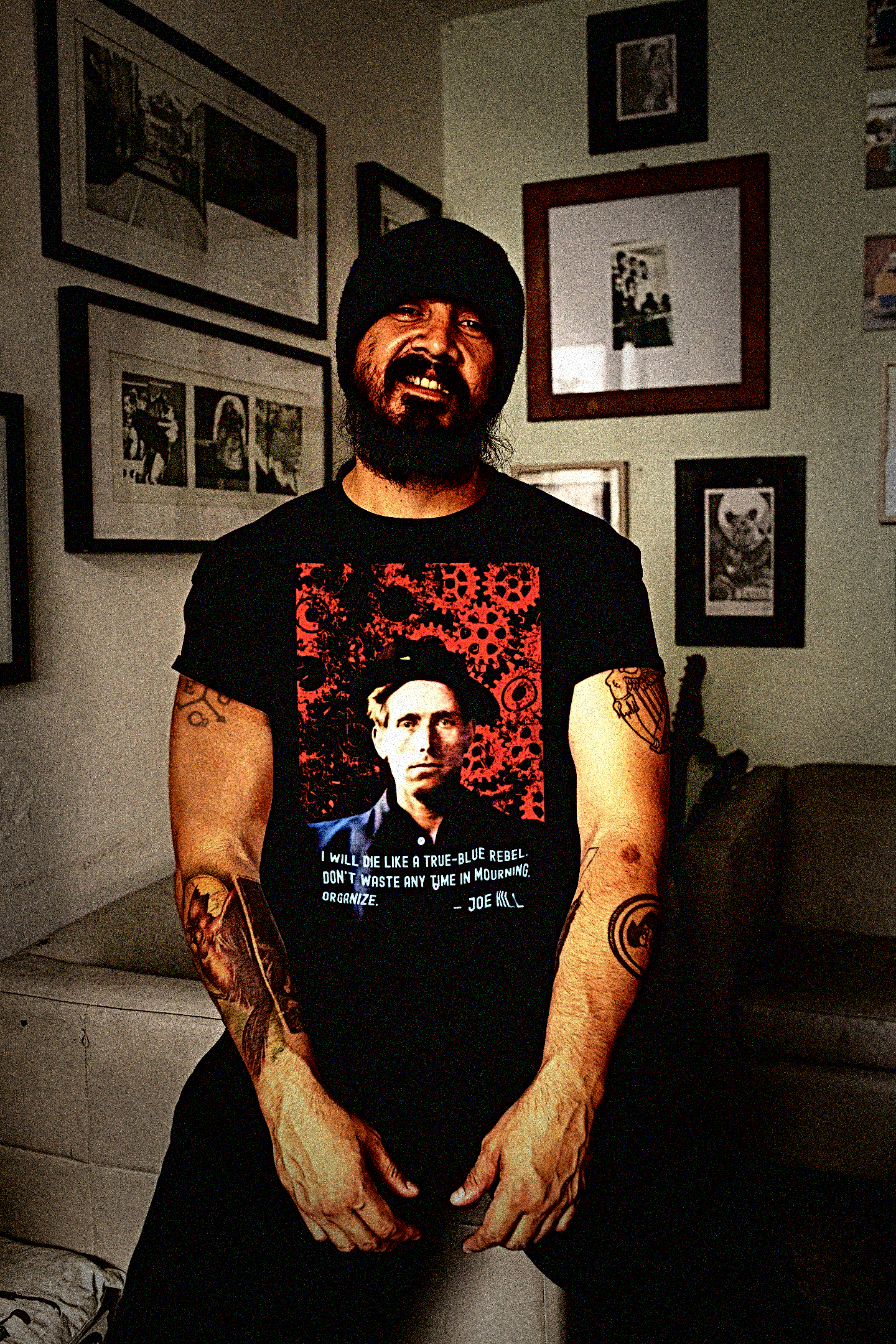 Tough bearded, tattooed Latino man wearing a Basic Black T-Shirt: Joe Hill, Wobbly IWW songwriter and labor martyr whose protest songs live on in union halls everywhere. “I will die like a true-blue rebel. Don't waste any time in mourning. Organize.”

Set in a living room with two salon walls of black and white photos.

Perfect gift for the labor organizer or anarcho-syndicalist in your life. Cotton Bella+Canvas 3001 #EUTC