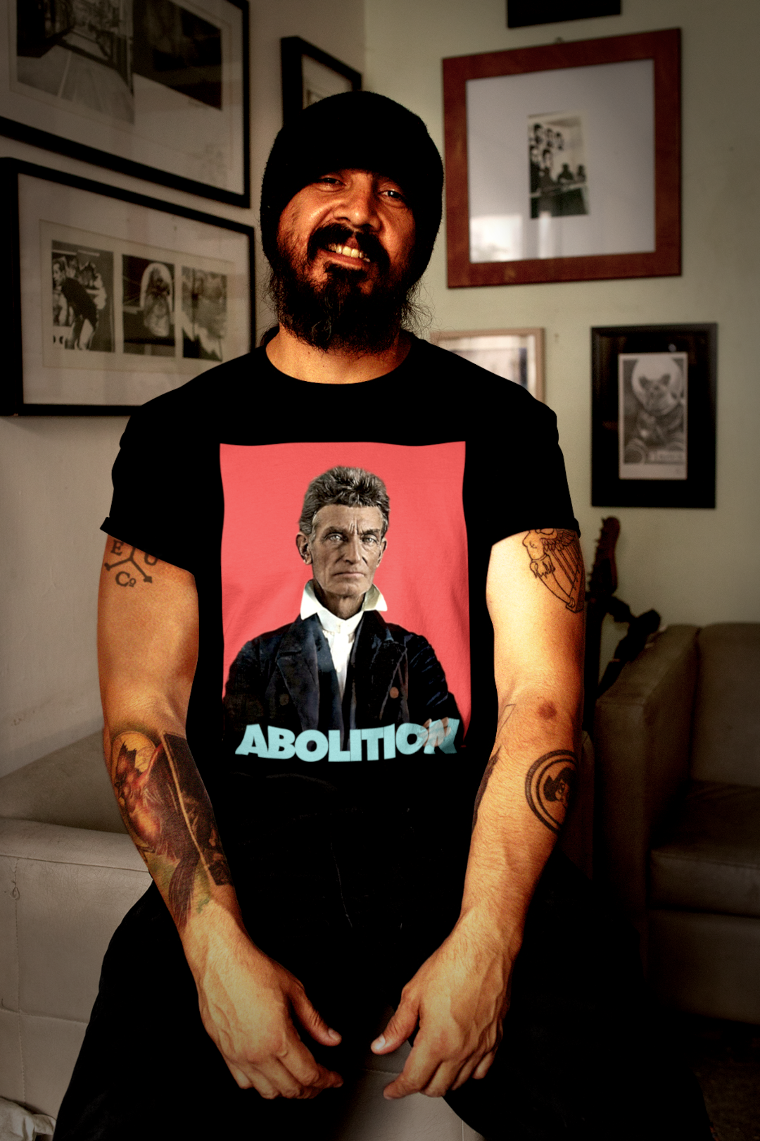 Bearded Tattooed Latino_Man_with Wool Cap in Living Room with Gallery Walls wearing Basic Black T-Shirt: John Brown, militant abolitionist who fought slavery with uncompromising zeal and sacrifice. Cotton Bella+Canvas 3001 #EUTC