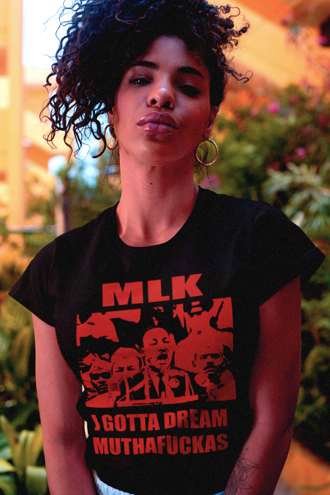 Beautiful Black woman with kinky hair updo  wearing a black relaxed fit women's t-shirt with red text and graphics featuring Martin Luther King Jr: I GOTTA A DREAM MUTHAFUCKAS in a tropical garden. 