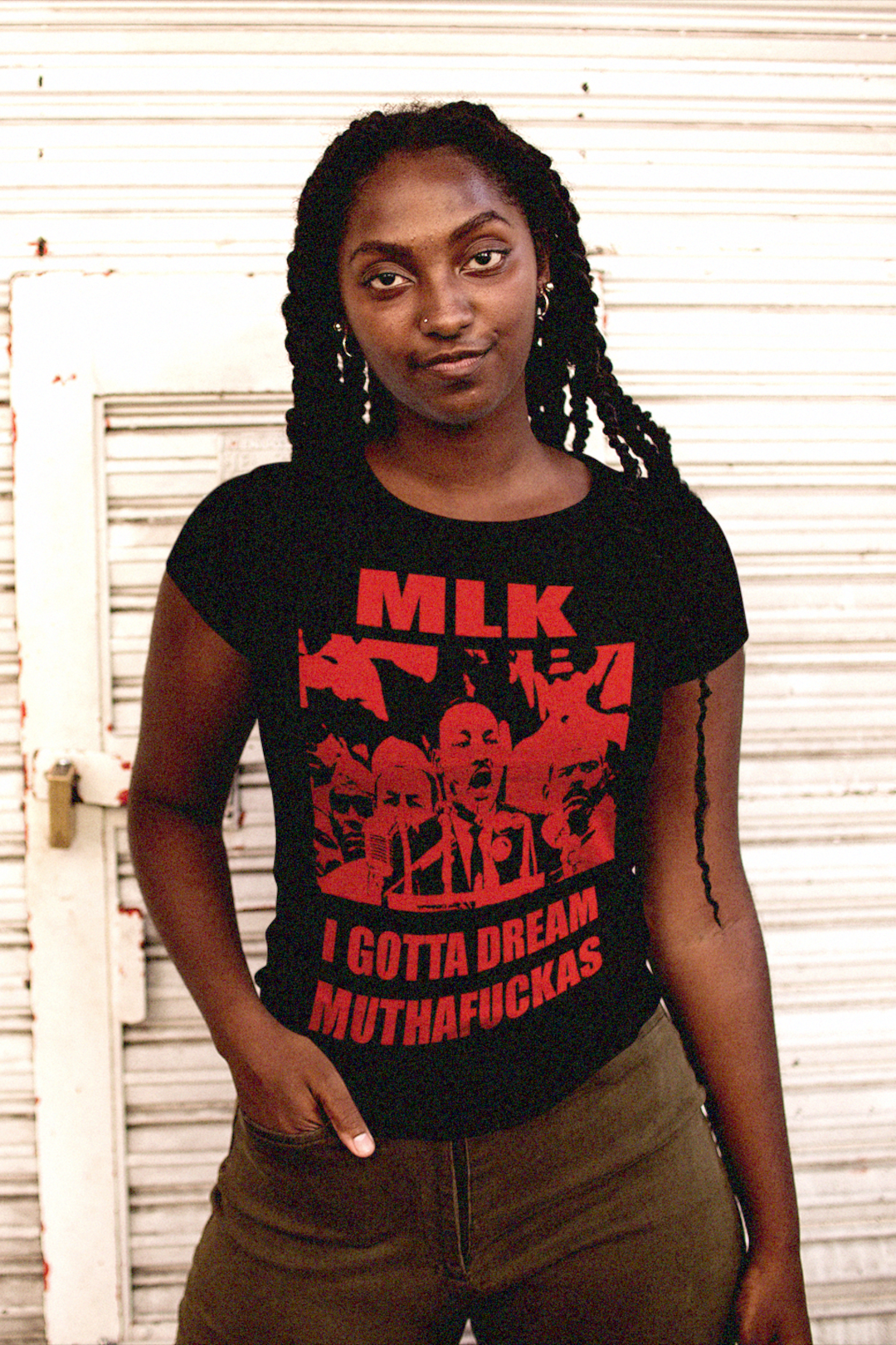 Strong Black woman with braided dreadlocks  wearing a black relaxed fit women's t-shirt with red text and graphics featuring Martin Luther King Jr: I GOTTA A DREAM MUTHAFUCKAS in front of a white industrial wall.