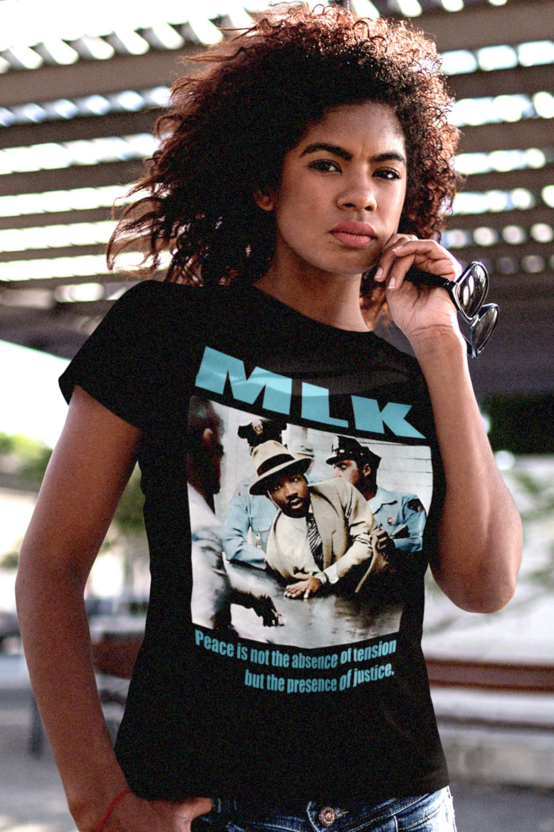 Strong Black woman wearing a black relaxed fit women's tee showing a colorized image of Martin Luther King Jr. Arrested in Montgomery (1956) with the quote: “Peace is not the absence of tension but the presence of justice.” Cotton shirt is the perfect gift for the civil rights activist in your life. Photo shows style and fit of tee. Only at #EUTC