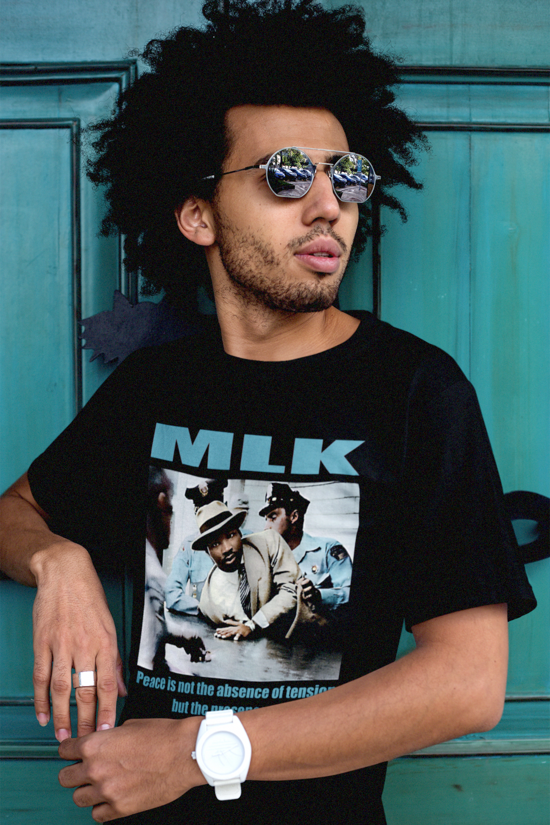 Cool man in reflector sunglasses and natural afro  wearing a Black Bella + Canvas 3001 Basic T-Shirt with a colorized photo of Martin Luther King's 1958 arrest in Memphis with the quote 'Peace is not the absence of tension but the presence of justice.' in bold teal letters. 

He is in front of a teal door. Photo to show style and fit of t-shirt. Perfect gift for the civil rights activist in your life. #EUTC