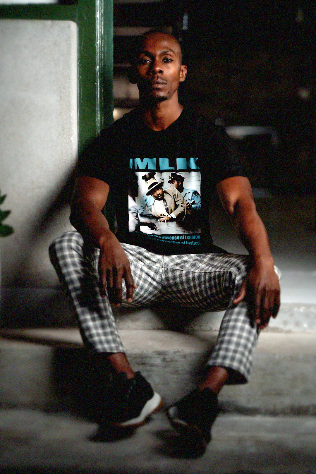 Cool man with close cropped hair wearing black and white plaid pants sitting on cement steps  wearing a Black Bella + Canvas 3001 Basic T-Shirt with a colorized photo of Martin Luther King's 1958 arrest in Memphis with the quote 'Peace is not the absence of tension but the presence of justice.' in bold teal letters. 

Perfect gift for the civil rights activist in your life. #EUTC