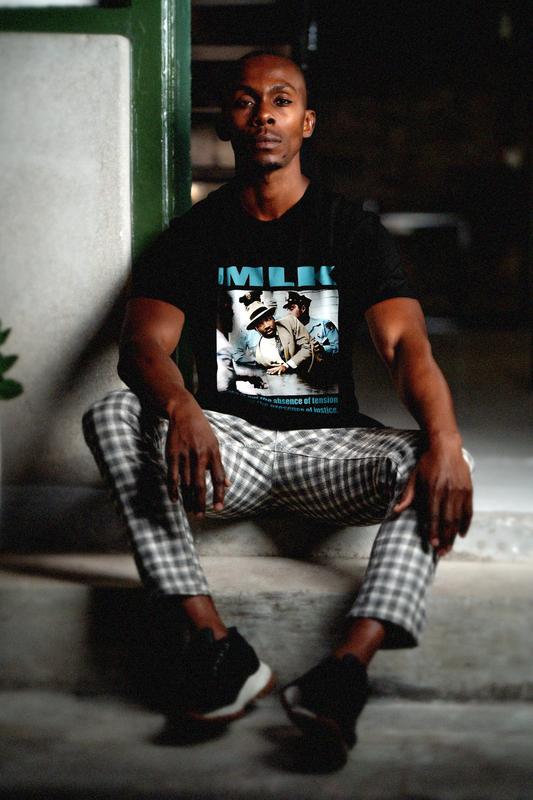 Cool man with close cropped hair wearing black and white plaid pants sitting on cement steps  wearing a Black Bella + Canvas 3001 Basic T-Shirt with a colorized photo of Martin Luther King's 1958 arrest in Memphis with the quote 'Peace is not the absence of tension but the presence of justice.' in bold teal letters. 

Perfect gift for the civil rights activist in your life. #EUTC