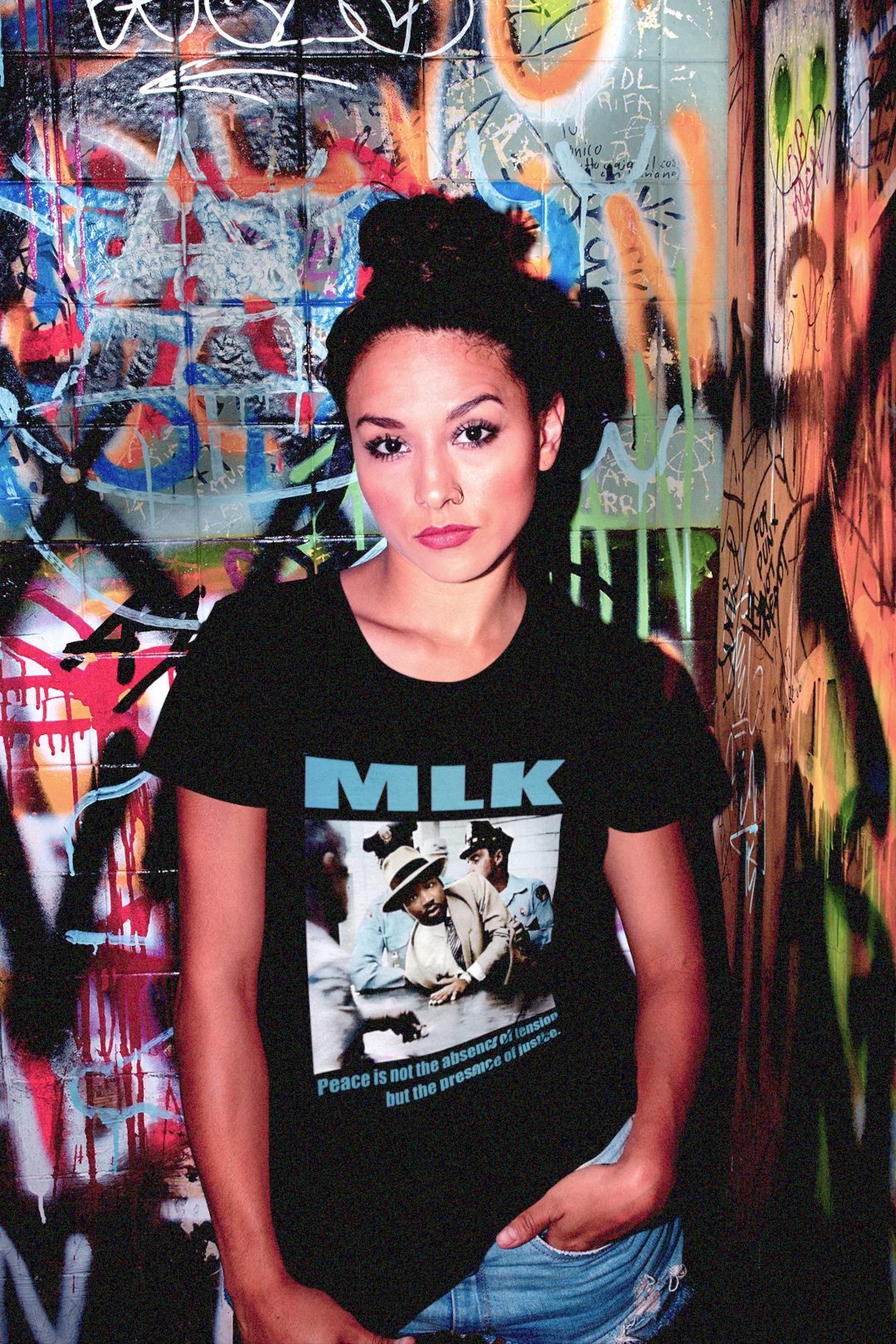Rebellious woman against grafitti cinder block wall wearing a black relaxed fit women's tee showing a colorized image of Martin Luther King Jr. Arrested in Montgomery (1956) with the quote: “Peace is not the absence of tension but the presence of justice.” Cotton shirt is the perfect gift for the rebel in your life. Photo shows style and fit of tee. Only at #EUTC