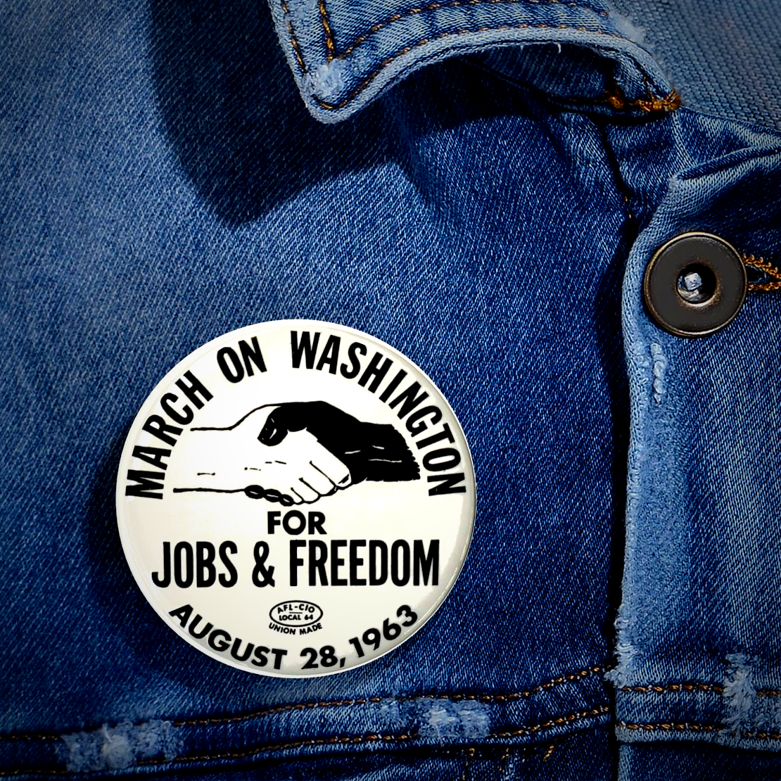A round white pin-back button with black text reading 'MARCH ON WASHINGTON FOR JOBS & FREEDOM AUGUST 28, 1963' and a handshake illustration, shown pinned to blue denim fabric.