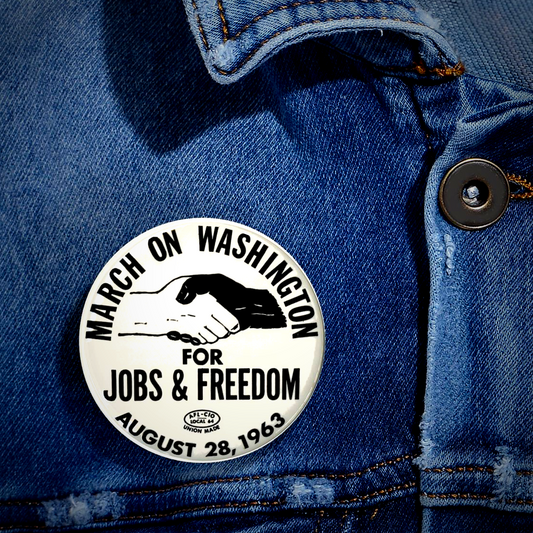 A round white pin-back button with black text reading 'MARCH ON WASHINGTON FOR JOBS & FREEDOM AUGUST 28, 1963' and a handshake illustration, shown pinned to blue denim fabric.