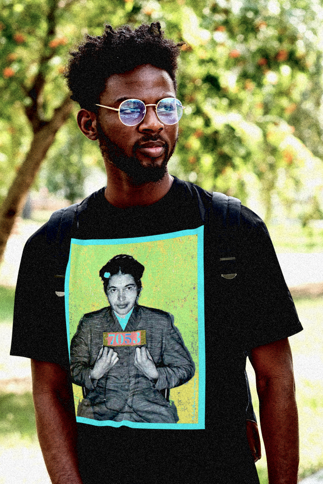 Conscious young Black man on campus with short dreadlocks,  Lennon glasses and a book bag wearing a basic black t-shirt depicting the mugshot of Rosa Parks, whose quiet defiance sparked the Montgomery Bus Boycott and galvanized the movement. Forget the myth of the tired seamstress—Rosa Parks was a trained activist who knew exactly what she was doing when she refused to give up her seat. Perfect gift for the feminist civil rights activist in your life. Bella+Canvas 3001 #EUTC