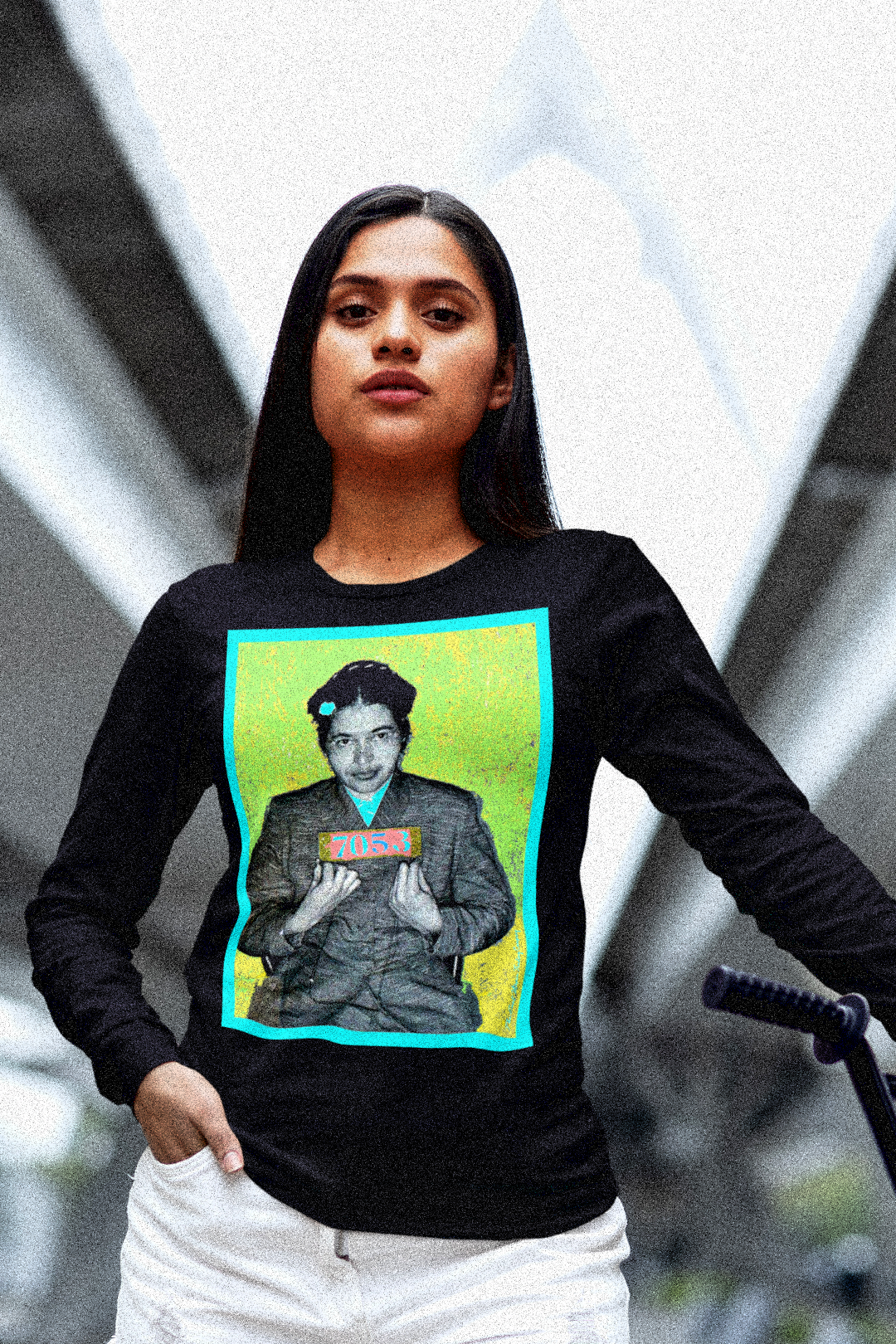 Young Latina woman with mountain bike in an urban area wearing a Black Long Sleeve Jersey depicting the mugshot of Rosa Parks, whose quiet defiance sparked the Montgomery Bus Boycott and galvanized the movement. Forget the myth of the tired seamstress—Rosa Parks was a trained activist who knew exactly what she was doing when she refused to give up her seat. Perfect gift for the feminist civil rights activist in your life. Bella+Canvas 3501  #EUTC 