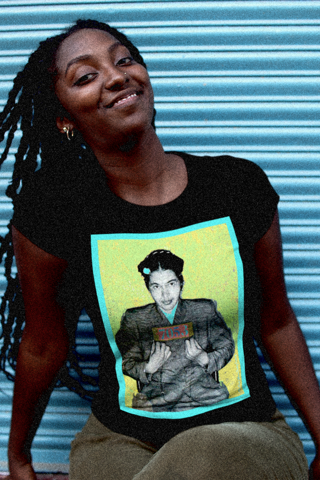 Strong Black woman with long dreadlocks in front of blue industrial door wearing a relaxed fit women's tee depicting the mugshot of Rosa Parks, whose quiet defiance sparked the Montgomery Bus Boycott and galvanized the movement. Forget the myth of the tired seamstress—Rosa Parks was a trained activist who knew exactly what she was doing when she refused to give up her seat. Perfect gift for the feminist civil rights activist in your life. Bella+Canvas Women’s Relaxed Tee 6400  #EUTC 