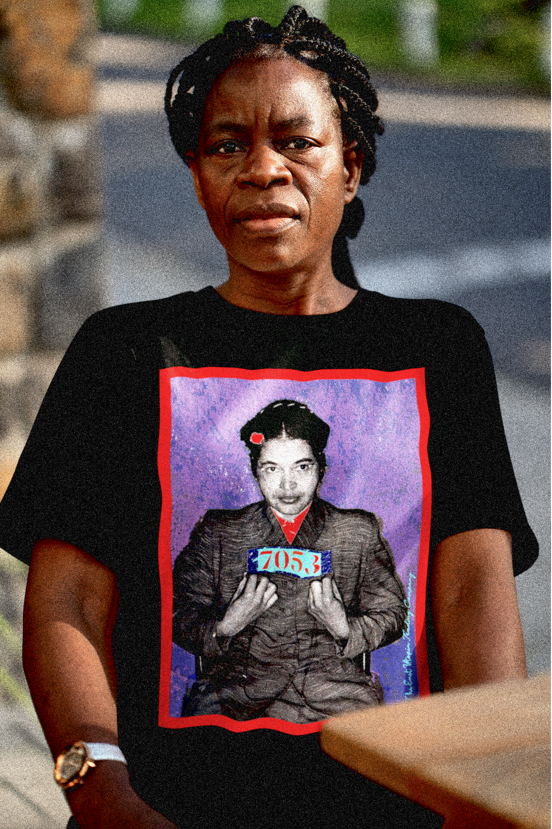 Strong, older Black woman with thick long braided hair pulled back sitting at a cafe table wearing a basic black t-shirt depicting the mugshot of Rosa Parks, whose quiet defiance sparked the Montgomery Bus Boycott and galvanized the movement. Forget the myth of the tired seamstress—Rosa Parks was a trained activist who knew exactly what she was doing when she refused to give up her seat. Perfect gift for the feminist civil rights activist in your life. Bella+Canvas 3001  #EUTC 