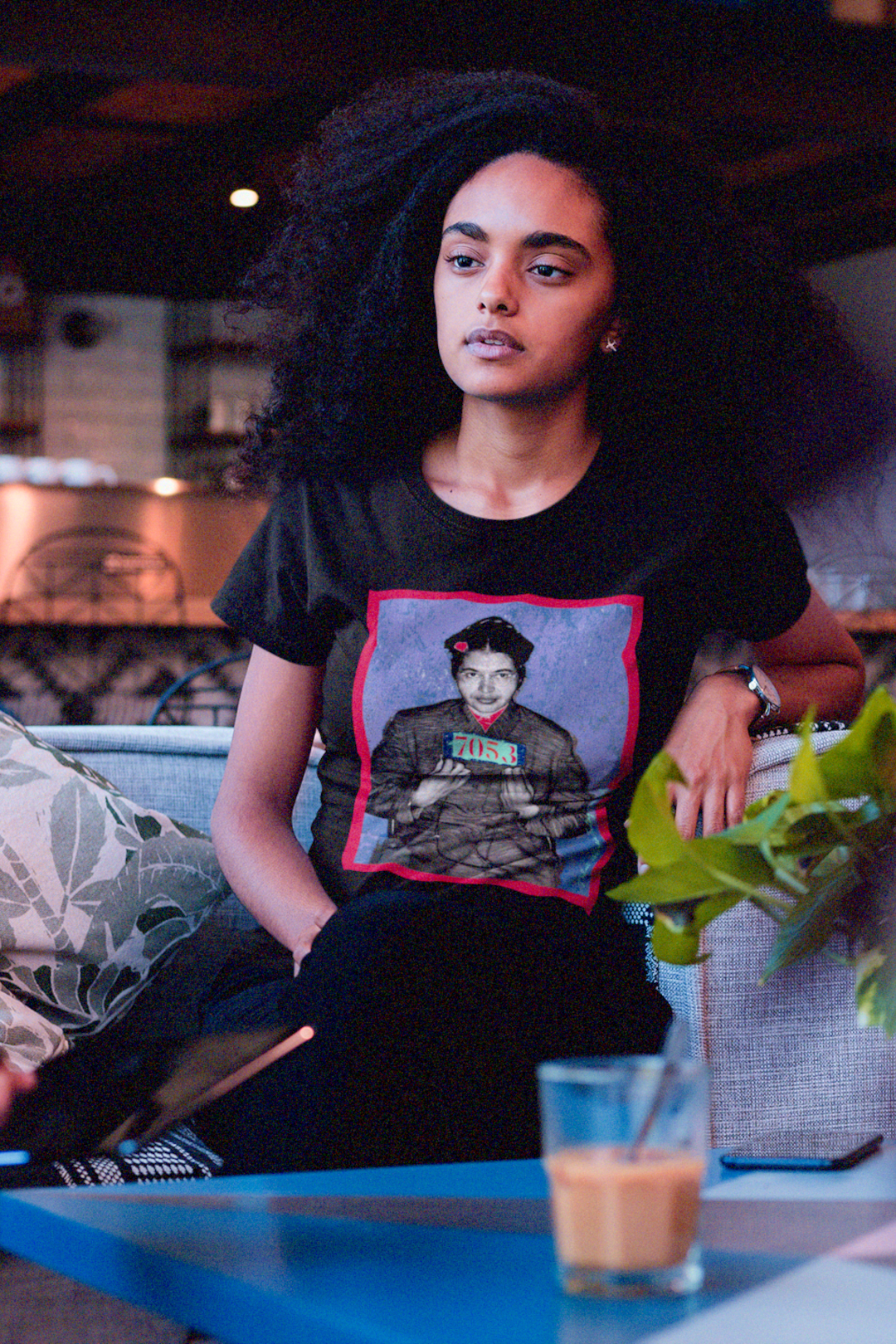 Young intellectual Black woman with large natural afro sitting at a cafe table wearing a basic black t-shirt depicting the mugshot of Rosa Parks, whose quiet defiance sparked the Montgomery Bus Boycott and galvanized the movement. Forget the myth of the tired seamstress—Rosa Parks was a trained activist who knew exactly what she was doing when she refused to give up her seat. Perfect gift for the feminist civil rights activist in your life. Bella+Canvas 3001  #EUTC 