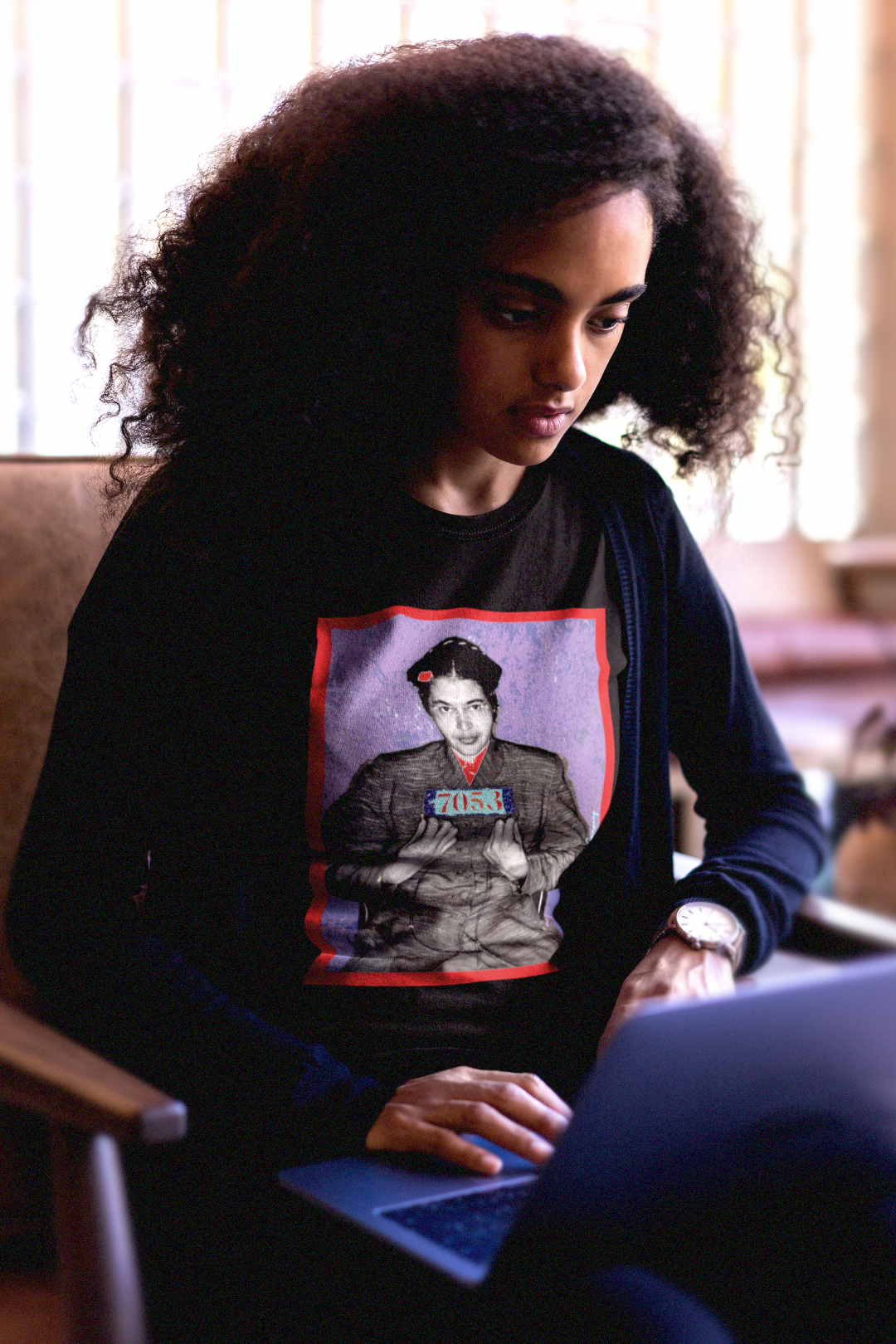 Intellectual Black woman with large natural afro in a college lounge working on a laptop wearing a Black Long Sleeve Jersey depicting the mugshot of Rosa Parks, whose quiet defiance sparked the Montgomery Bus Boycott and galvanized the movement. Forget the myth of the tired seamstress—Rosa Parks was a trained activist who knew exactly what she was doing when she refused to give up her seat. Perfect gift for the feminist civil rights activist in your life. Bella+Canvas 3501  #EUTC
