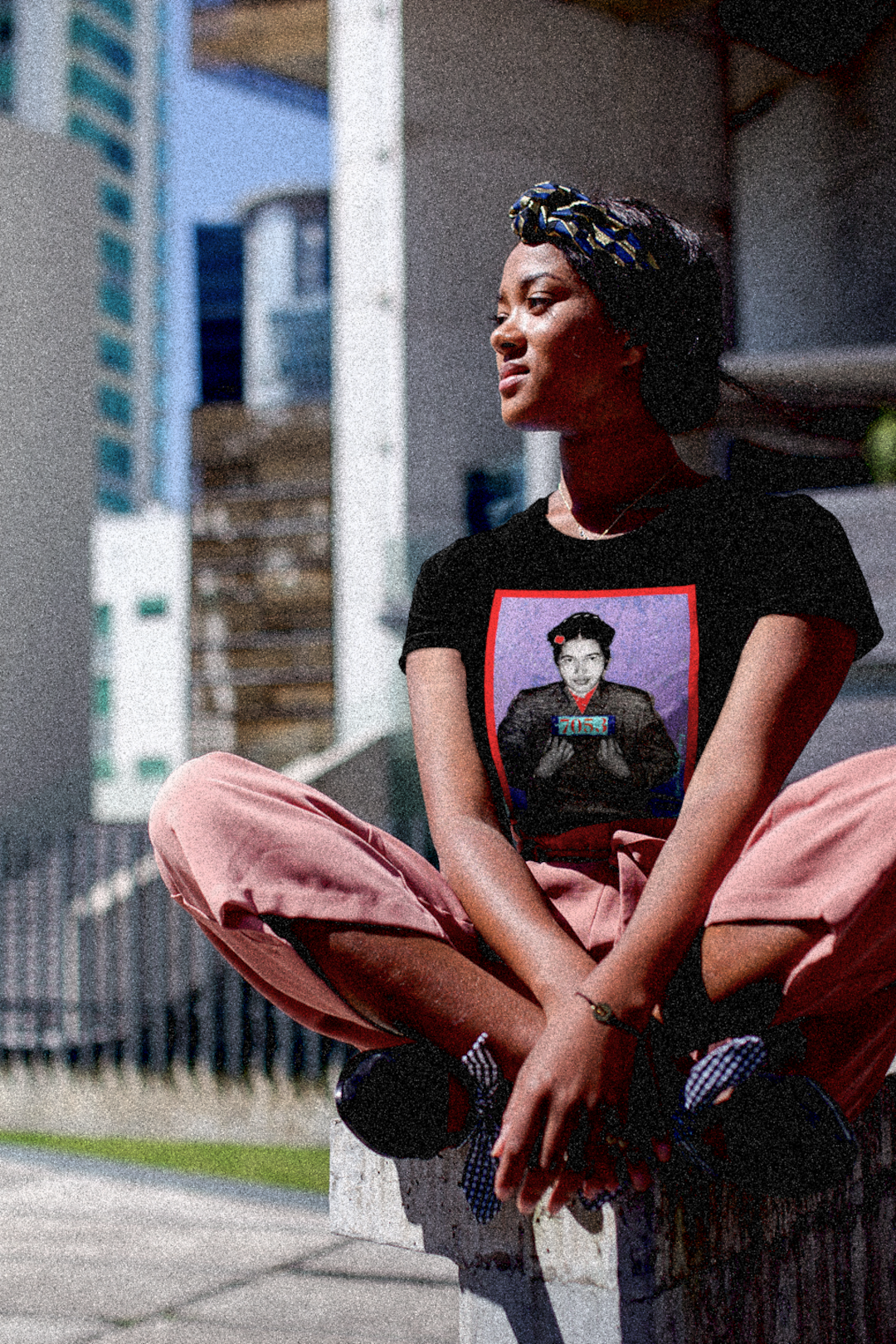 Strong Black Woman with Stylish Head Scarf sitting Cross-legged in City Square wearing a relaxed fit women's tee depicting the mugshot of Rosa Parks, whose quiet defiance sparked the Montgomery Bus Boycott and galvanized the movement. Forget the myth of the tired seamstress—Rosa Parks was a trained activist who knew exactly what she was doing when she refused to give up her seat. Perfect gift for the feminist civil rights activist in your life. Bella+Canvas Women’s Relaxed Tee 6400 #EUTC 