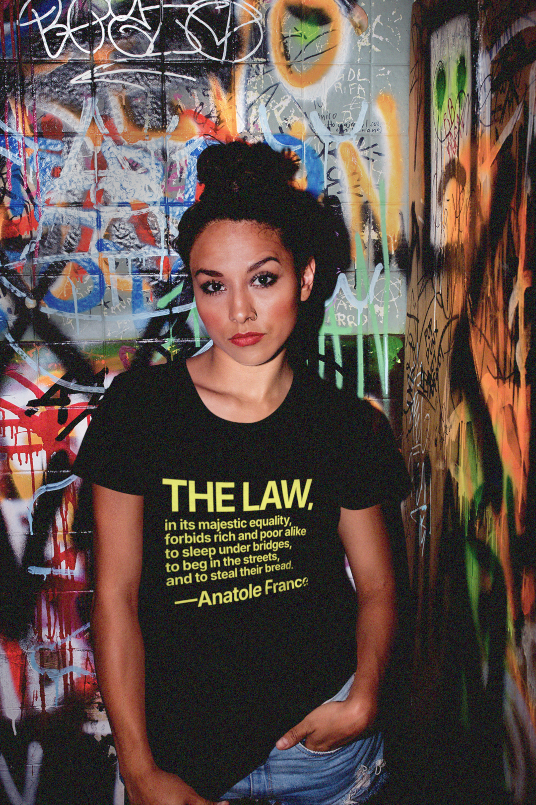 Woman wearing a black relaxed fit t-shirt with yellow text in front of a graffiti-covered night club wall - Anatole France quote: “The law, in its majestic equality, forbids rich and poor alike to sleep under bridges, to beg in the streets, and to steal their bread.” 