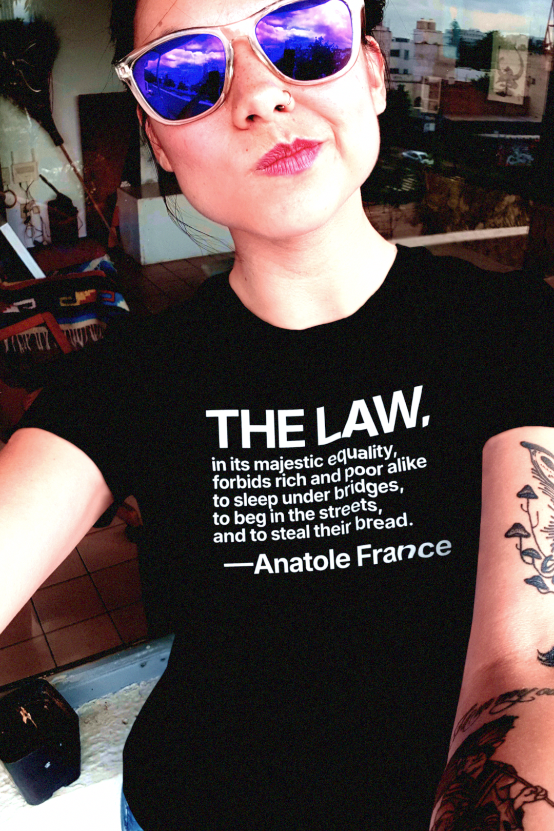 Woman wearing a black relaxed fit t-shirt with a quote and sunglasses indoors. Anatole France quote: “The law, in its majestic equality, forbids rich and poor alike to sleep under bridges, to beg in the streets, and to steal their bread.” 