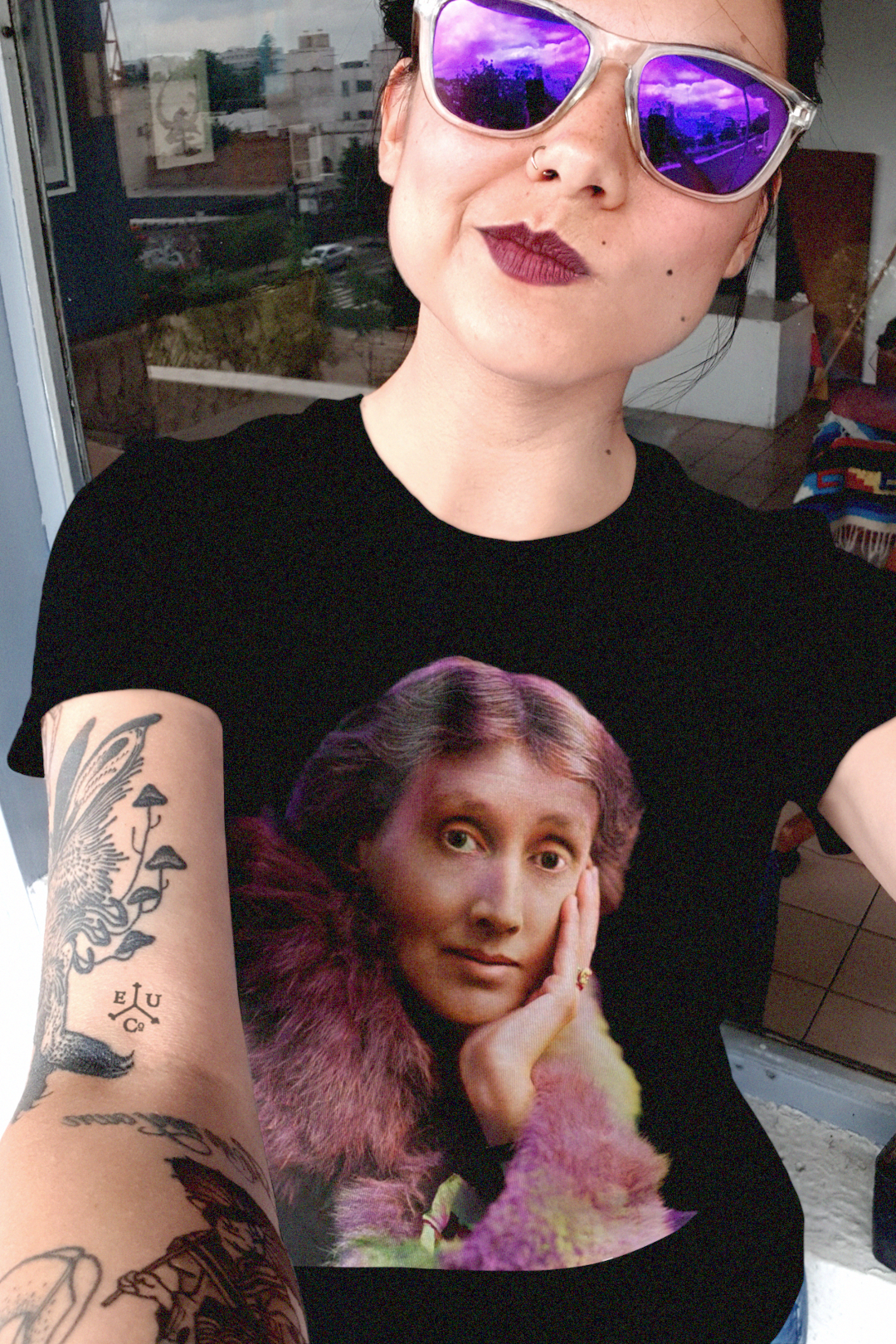 Tattooed woman with vintage framed purple reflector sunglasses wearing a relaxed fit women's tee of a technicolor Virginia Woolf. She made literature new—stream-of-consciousness, fractured time, inner lives spilling across the page. Quality cotton tee. Perfect gift for the lover of literature in your life. Bella+Canvas Women’s Relaxed Tee 6400 #EUTC