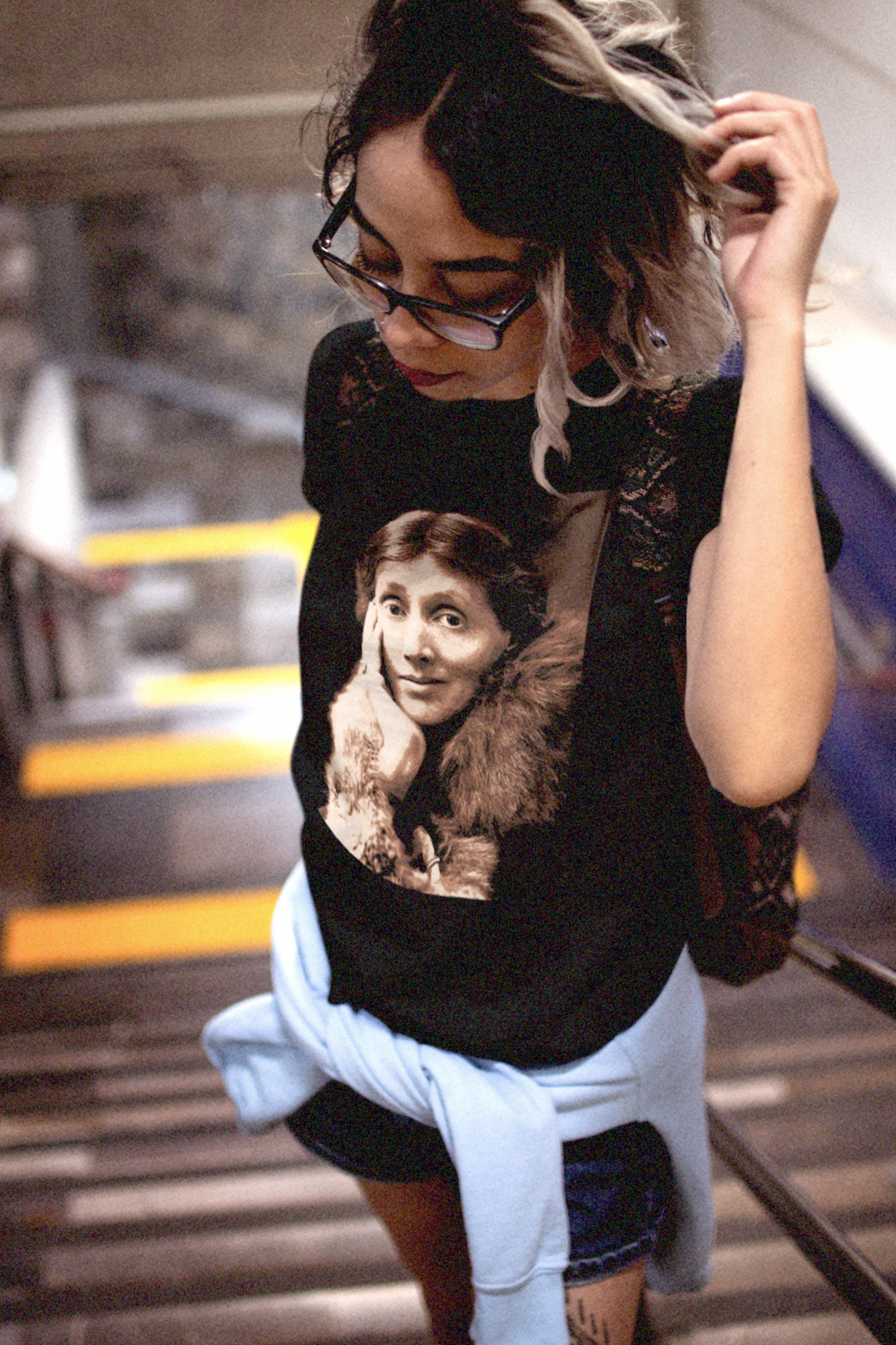 Emo Latina with a funky bookbag heavy framed glasses on a subway stairway wearing a relaxed fit women's Tee with a sepia portrait of Virginia Woolf. She made literature new—stream-of-consciousness, fractured time, inner lives spilling across the page. With novels like Mrs. Dalloway and To the Lighthouse, she redefined modernism not as escape but as immersion. Photo show fit and style. Perfect gift for your book club fit or the English major in your life. #EUTC