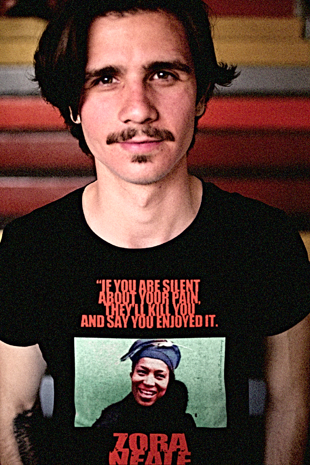 Zora Neale Hurston - Basic T-Shirt - White guy with van dyke mustache and beard on campus in front of colorful stairs wearing a basic Black t-shirt depicting Zora Neale Hurston: If you are silent about your pain, they'll kill you and say you enjoyed it. Zora Neale Hurston didn’t just write novels; she bottled the rhythm and brilliance of Black life in the South and poured it straight onto the page. Cotton Bella+Canvas 3001  #EUTC